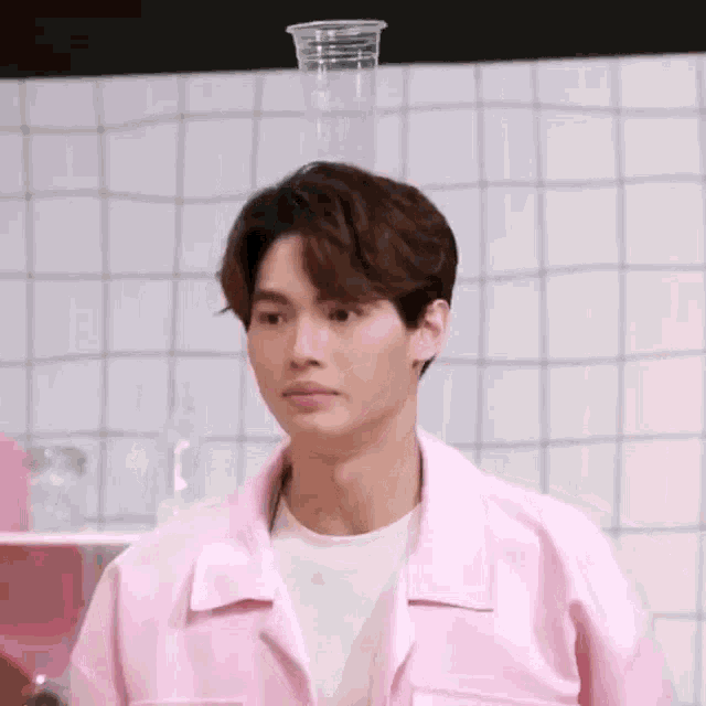 a young man in a pink jacket is standing in front of a white tiled wall with a plastic cup on his head .