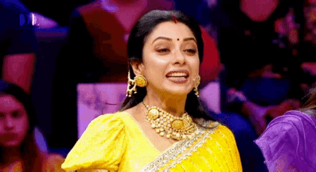 a woman in a yellow saree and gold jewelry is smiling while standing in front of a crowd .