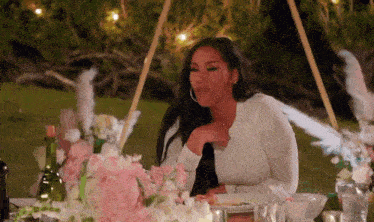 a woman in a white shirt is sitting at a table with flowers and a plate of food .