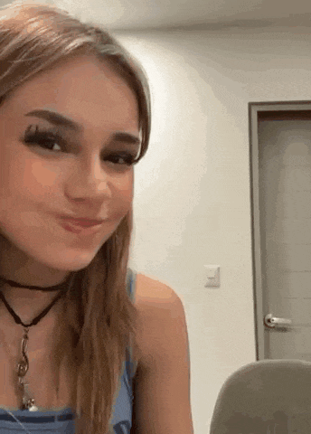 a young woman wearing a choker and a blue tank top is sitting in a chair and smiling .