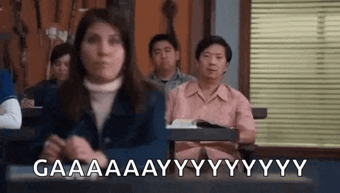 a woman is sitting at a desk in a classroom with other people and making a funny face .