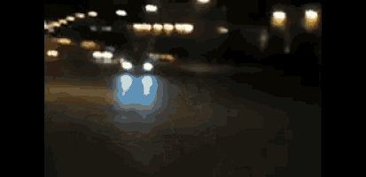 a group of people are standing in front of a car that is driving down the street at night
