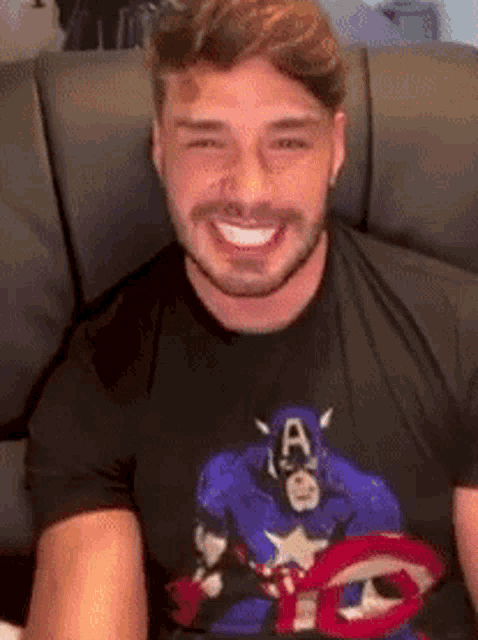 a man wearing a captain america t-shirt is smiling while sitting on a couch .