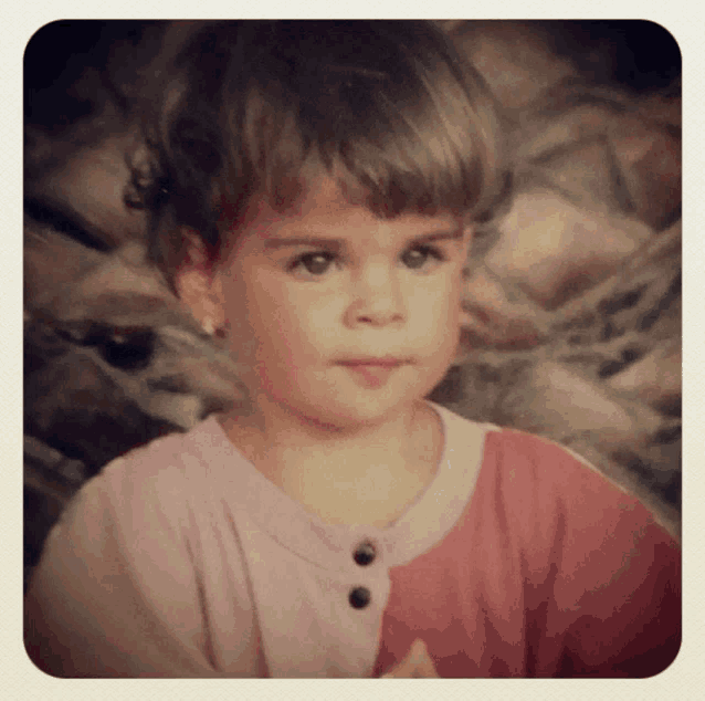a little girl wearing a pink shirt with black buttons looks at the camera