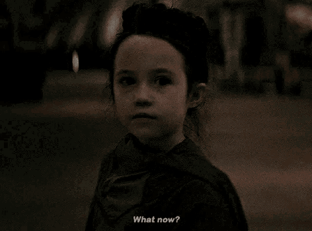 a little girl is standing in the dark talking to someone and asking what now .