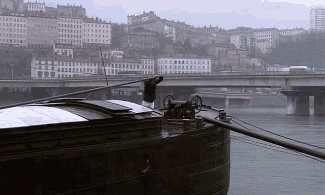 a man stands on the deck of a boat with a bridge in the background and a building that says ford on it