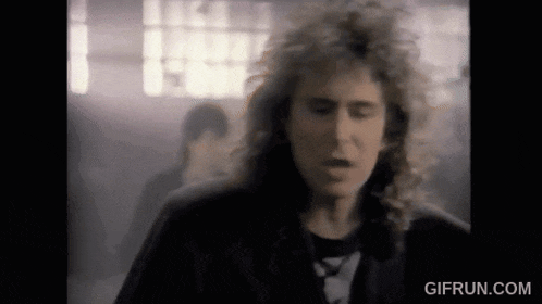 a man with long curly hair is standing in a dark room with smoke coming out of his mouth .