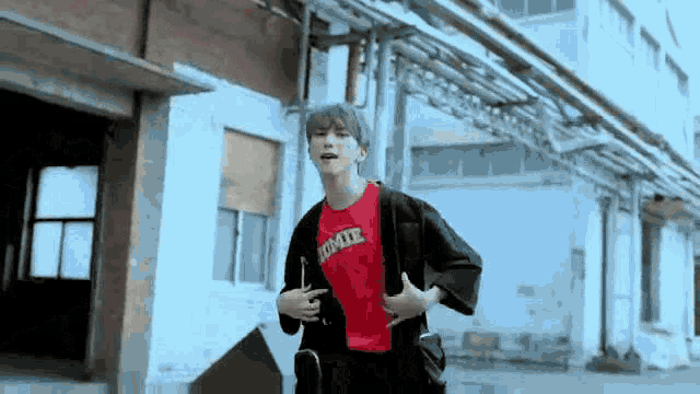 a young man is standing in front of a building wearing a red t-shirt and a black jacket .