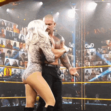a woman in a bodysuit is hugging a man in a wrestling ring with the word choccy wrestling on the wall