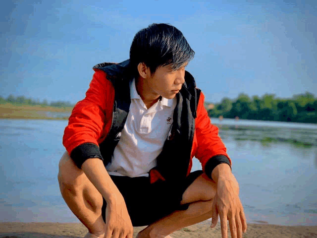 a man wearing a red jacket and a white polo shirt is squatting on the beach