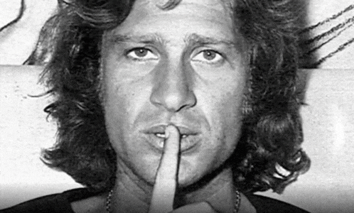 a man with long hair is holding his finger to his mouth and making a silence sign .