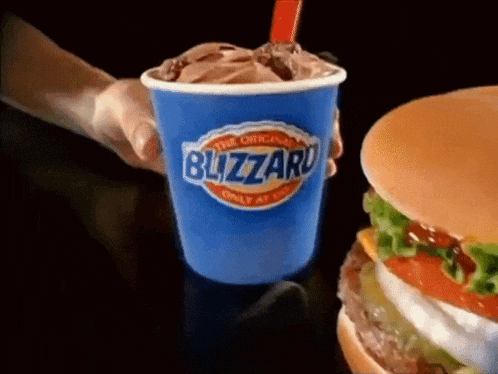 a person is holding a cup of blizzard ice cream in front of a hamburger