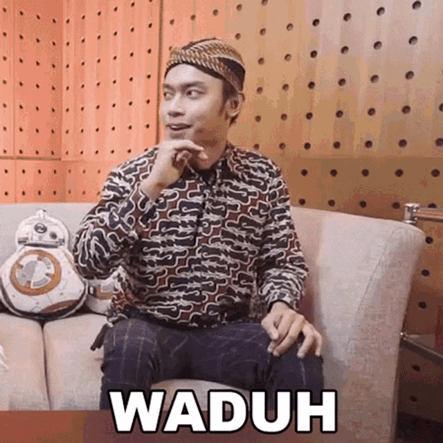a man wearing a hat is sitting on a couch with the word waduh written on the bottom .