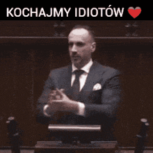 a man in a suit and tie is clapping his hands with the words kochajmy idiotow above him .