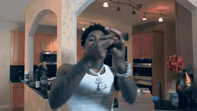 a man wearing a white tank top and a necklace with the letter s on it is standing in a kitchen