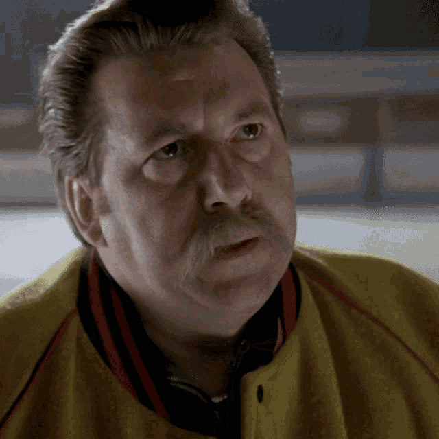 a man with a mustache and a yellow jacket looks at the camera