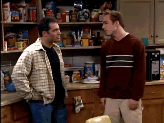two men are standing in a kitchen talking to each other and one of them is wearing a red sweater with stripes
