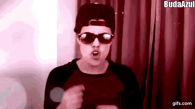 a man wearing sunglasses , a hat and a black shirt is making a funny face .