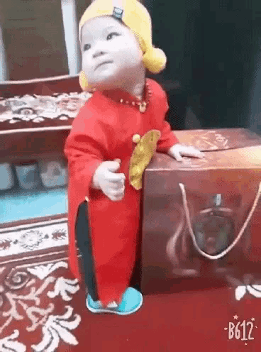 a baby wearing a red outfit and a yellow hat is standing next to a box on a rug .