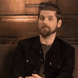 a man with a beard is sitting on a couch with a glass of beer in his hand .