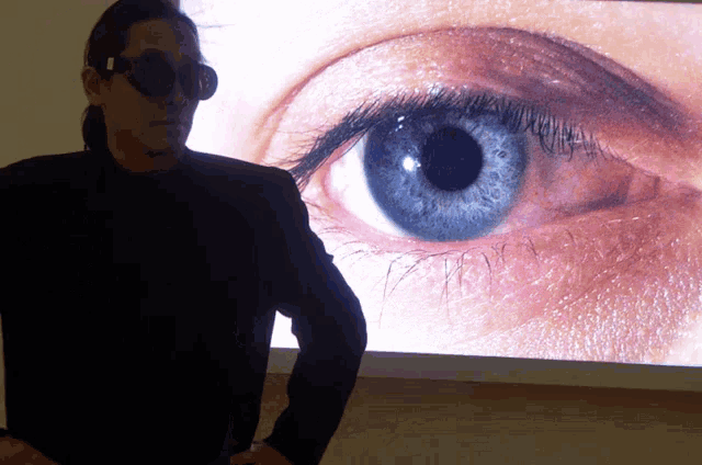a man wearing sunglasses stands in front of a large screen with a woman 's eye on it
