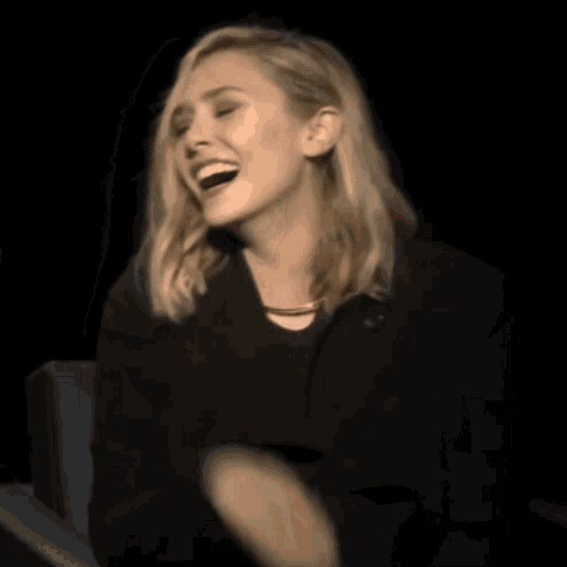 a woman in a black jacket is laughing in a dark room while sitting in a chair .