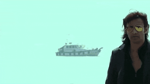 a man in a leather jacket and sunglasses is standing in front of a boat in the water .