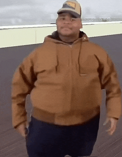 a man wearing a brown jacket and a hat is standing on a deck .