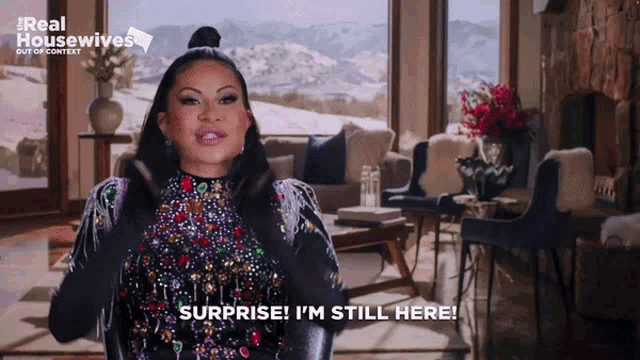 a woman says surprise i 'm still here while standing in a living room