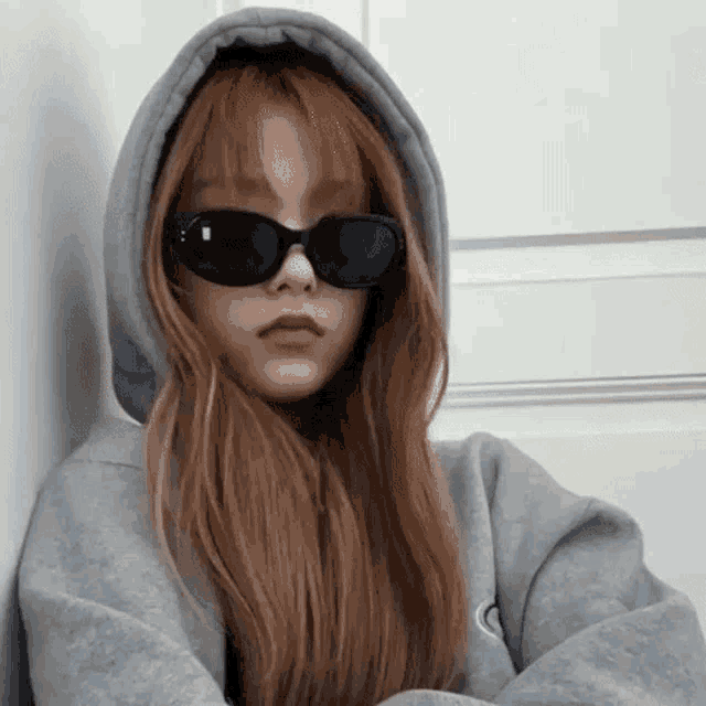 the girl is wearing a hoodie and sunglasses and leaning against a wall .
