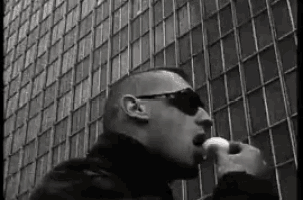 a man wearing sunglasses is eating a sandwich in front of a building in a black and white photo .