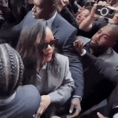 a woman wearing sunglasses and a suit is surrounded by a crowd of people .