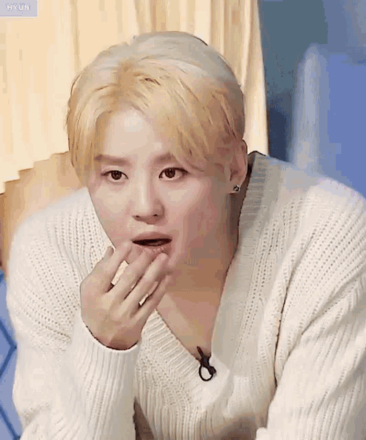 a young man with blonde hair is wearing a white sweater and covering his mouth with his hands .