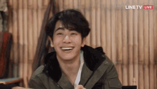 a young man in a green jacket is smiling while sitting at a table in front of a bamboo wall .