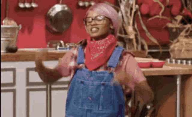 a woman wearing overalls and a bandana is standing in a kitchen .