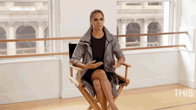 a woman sits in a director 's chair in front of a window with cosmopolitan.com in the corner
