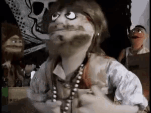 a puppet with a beard and a pearl necklace is standing in front of a skull and two other puppets .