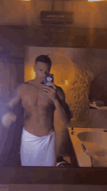 a shirtless man wrapped in a white towel takes a selfie with his phone