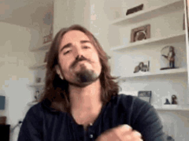 a man with long hair and a beard is making a funny face in front of a shelf .