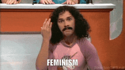 a man with curly hair and a mustache is wearing a pink shirt with a unicorn on it and says feminism
