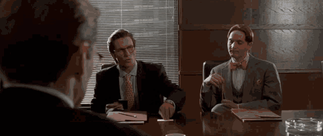 two men in suits and bow ties are sitting around a table
