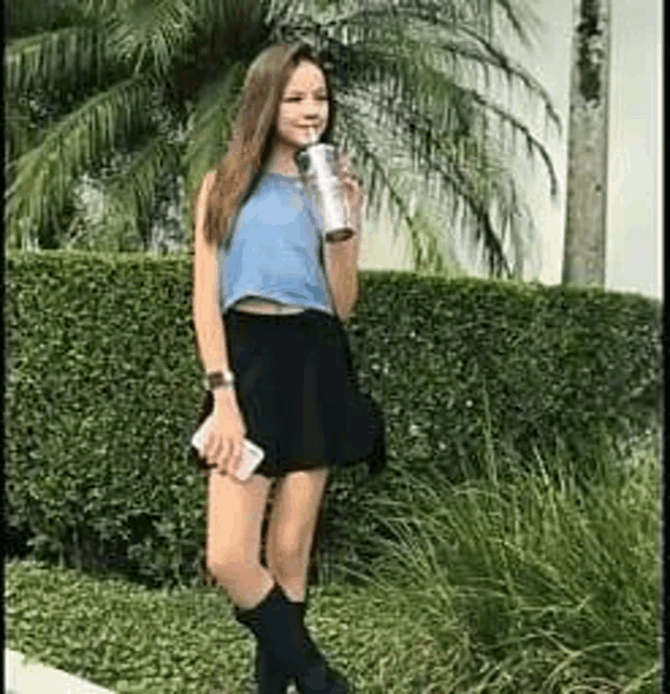 a girl is standing in front of a hedge holding a cup of coffee .
