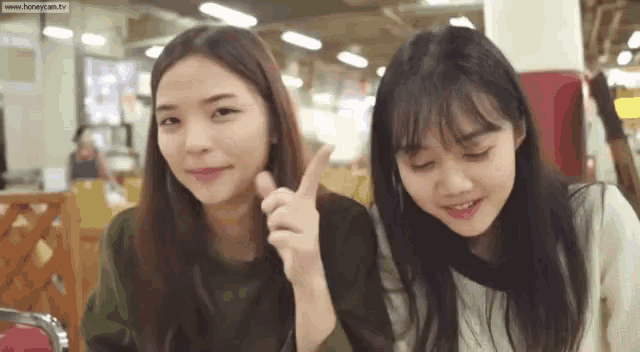 two girls are sitting next to each other in a restaurant and one of them is pointing at the camera .