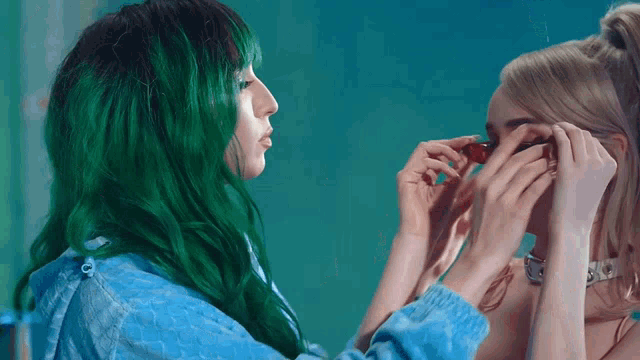 a woman with green hair adjusts a pair of sunglasses on another woman