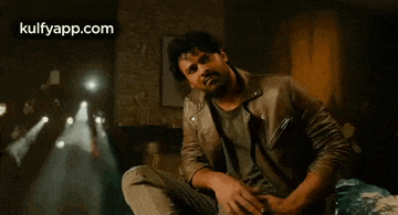 a man in a leather jacket is sitting on a couch in a dark room .