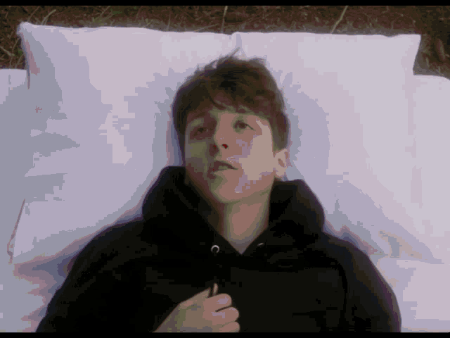 a young man in a black hoodie is laying on a bed with white pillows