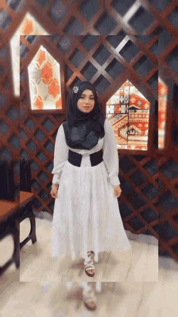 a woman in a white dress and black hijab is standing in front of a wooden wall