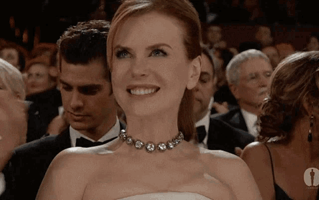 a woman in a white dress and a necklace smiles in front of an oscars logo