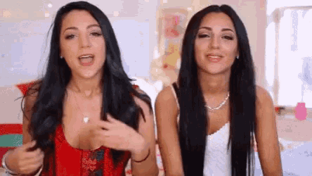 two women with long black hair are sitting next to each other in a bedroom .