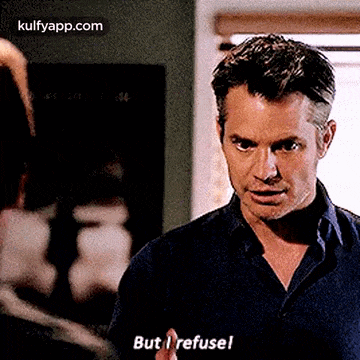 a man in a blue shirt is standing in front of a window and saying `` but i refuse ! ''